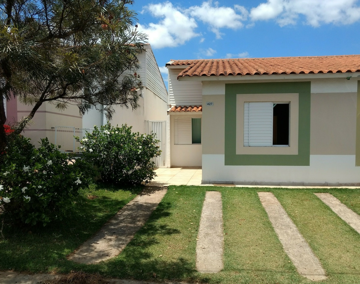 A single-story house is framed by greenery, featuring a terracotta roof and light-colored exterior walls with decorative green accents. A paved pathway leads to the entrance, which is complemented by a small yard with grass and shrubs, offering a sense of openness and accessibility.