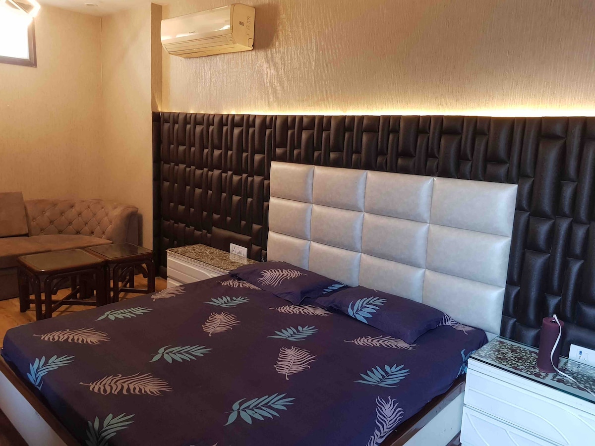 A spacious sleeping area features a large bed dressed in linens with a leaf pattern. Dark accent walls provide contrast to the light-colored bed. A comfortable seating area is visible in the corner, next to a coffee table, with an air conditioning unit mounted above.