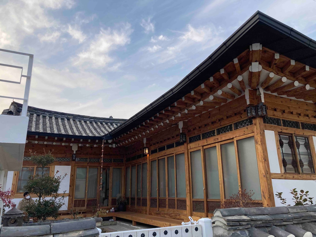 The exterior showcases a traditional hanok structure featuring wooden beams and open windows, complemented by a tiled roof. A small garden area with plants is visible, enhancing the natural surroundings. The overall design reflects a harmonious blend of classic and modern elements.