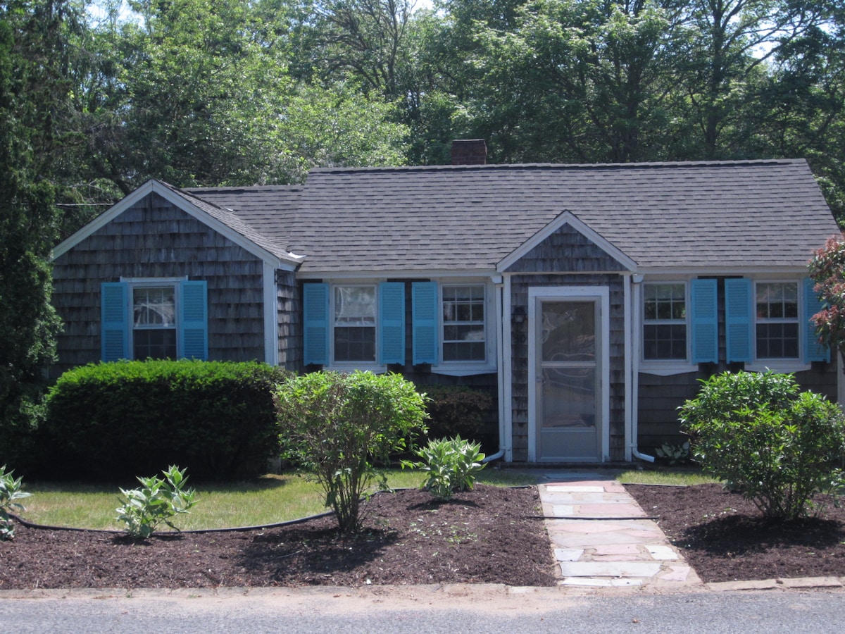 A charming Cape Cod cottage is shown featuring dark wooden shingles and bright blue shutters. The front porch is adorned with a screen door, and surrounded by landscaped shrubs and a stone pathway leading to the entrance, inviting guests into a cozy retreat.
