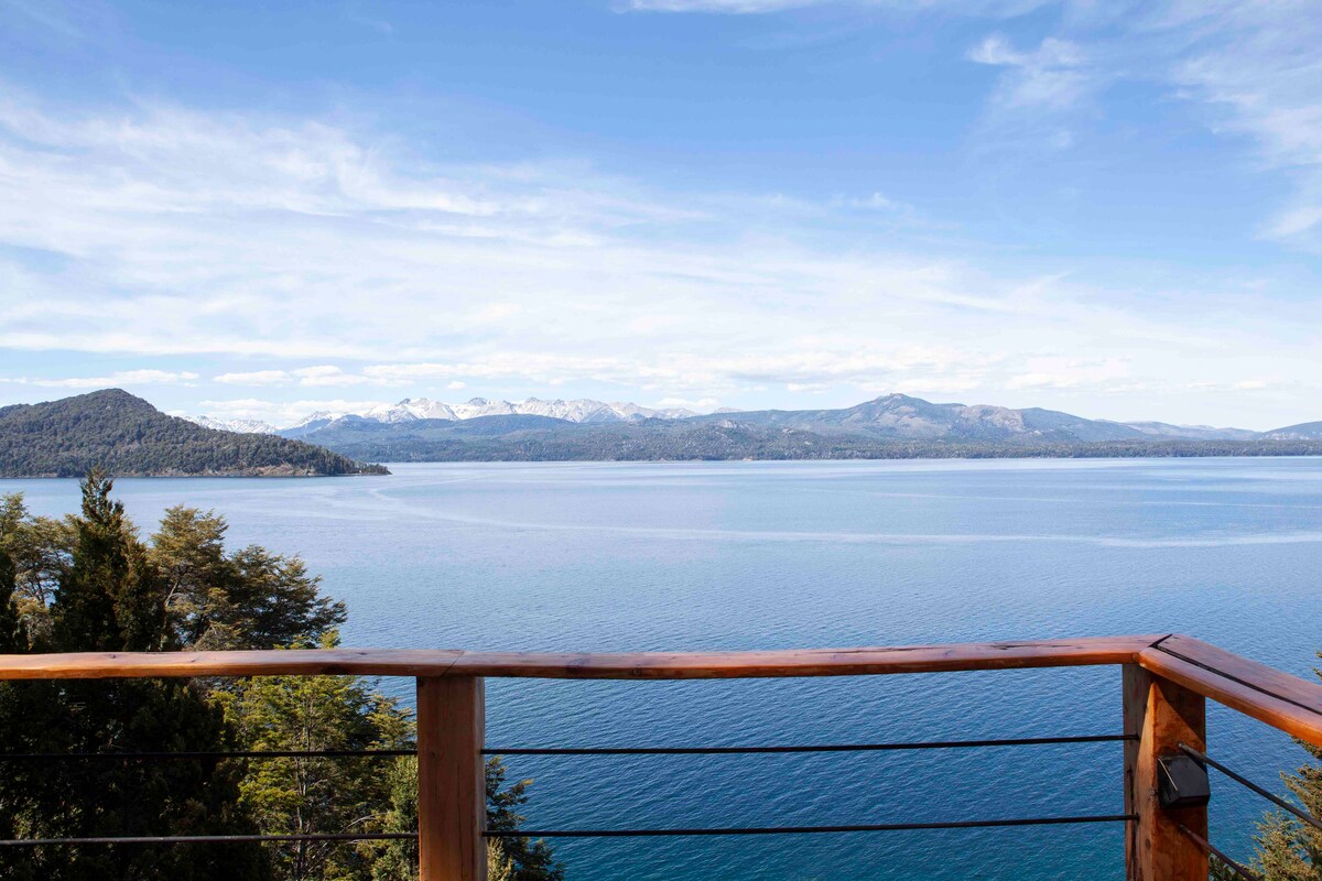 A serene view from the balcony terrace overlooks the expansive lake, with a backdrop of rugged mountains and a clear blue sky. The calm water reflects the landscape, creating a peaceful outdoor environment.