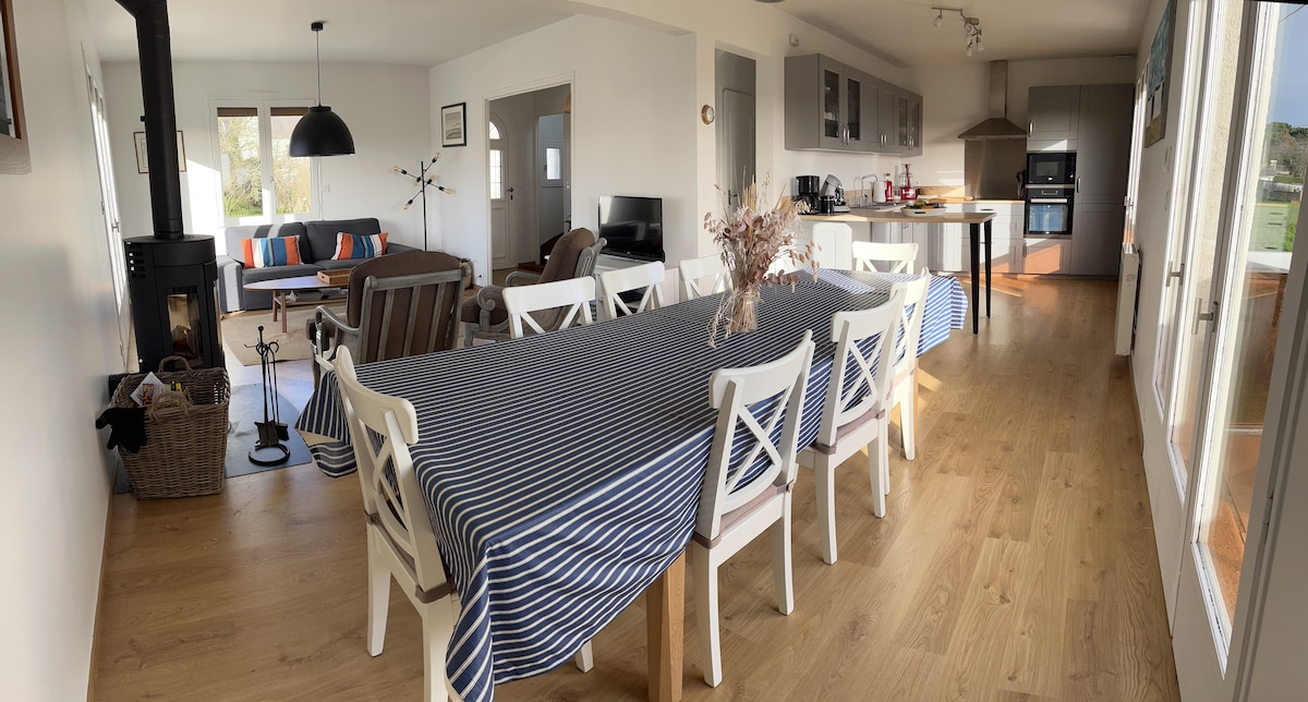 A spacious dining area features a long table with a striped blue and white tablecloth, surrounded by white chairs. A cozy seating area with a sofa is visible in the background, along with a wood stove. Natural light fills the room, highlighting the wooden flooring.