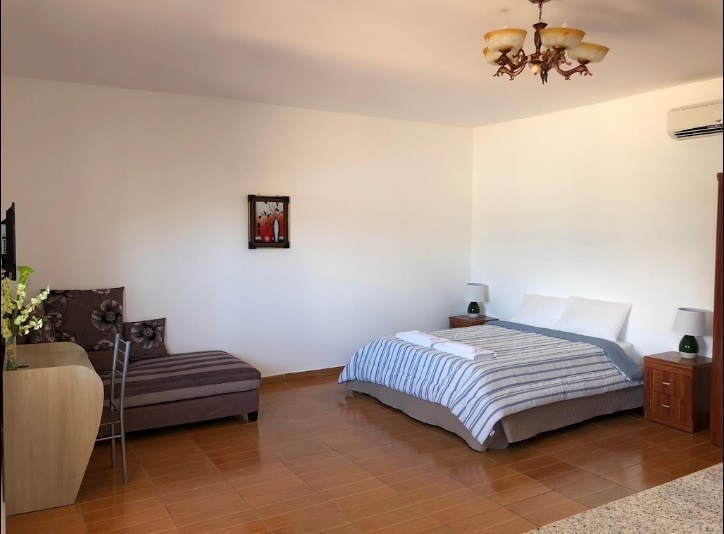 A spacious guest room features a comfortable bed with light, striped linens. A simple chair is positioned nearby, and a television is mounted on the wall. Warm light is provided by bedside lamps, and a decorative piece adds a touch of style to the room.