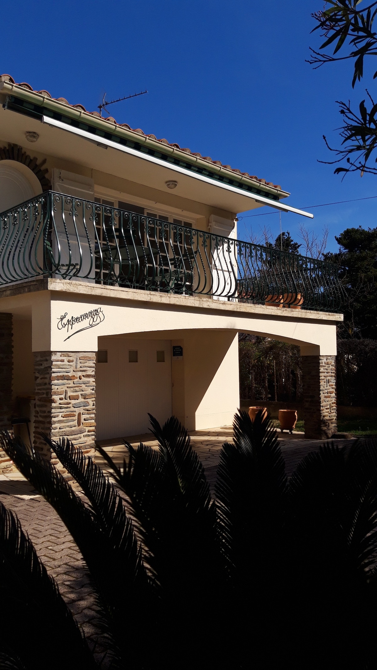 The exterior of the villa is displayed, highlighting a two-story structure with a balcony bordered by a decorative railing. Stone accents complement the building's façade, and the surrounding greenery adds a natural touch. Clear blue skies provide a bright backdrop.