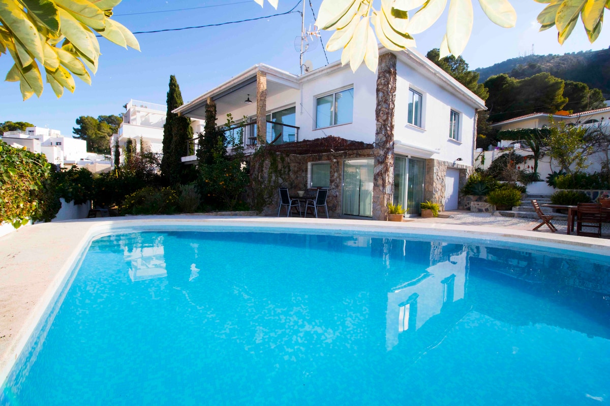 A renovated two-story villa is seen from the pool area, showcasing its modern architecture and large windows. The vibrant blue pool reflects sunlight, surrounded by lush greenery and patio chairs. The exterior features natural stone accents and a spacious outdoor area for relaxation.