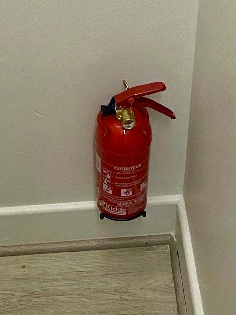 A red fire extinguisher is mounted on a wall in a corner of the room. The device is positioned near the floor, featuring a clear safety pin and handle, indicating readiness for use in emergencies.