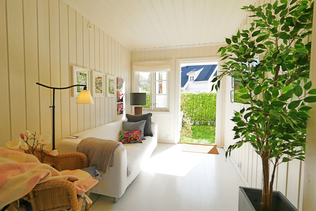 A bright and airy living area features a white sofa adorned with colorful cushions. Natural light filters in through a window, illuminating a houseplant and woven chair. Framed artwork is displayed on the walls, enhancing the inviting atmosphere of the space.