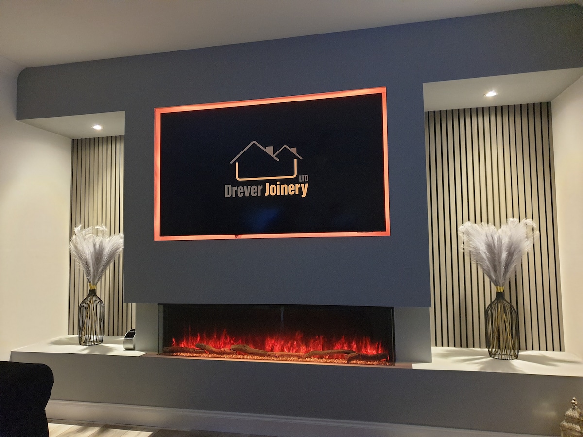A sleek entertainment wall features a modern electric fireplace with a glowing reddish hue. Two vases hold soft, feathery plants, flanking a centrally mounted flat-screen television. The understated color palette complements the vertical wooden paneling behind, creating a contemporary look for relaxation or socializing.