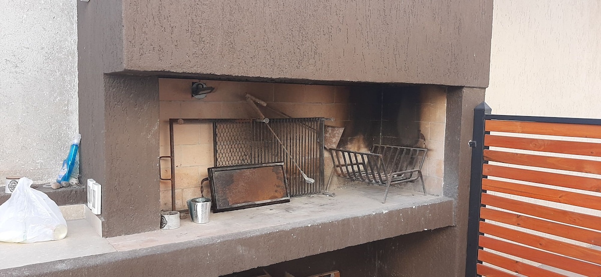 An outdoor barbecue area is featured, with a built-in stone structure housing a grilling space and a metal rack. A large grate is visible alongside a flat surface below, and the area is enclosed by a wooden fence for privacy.