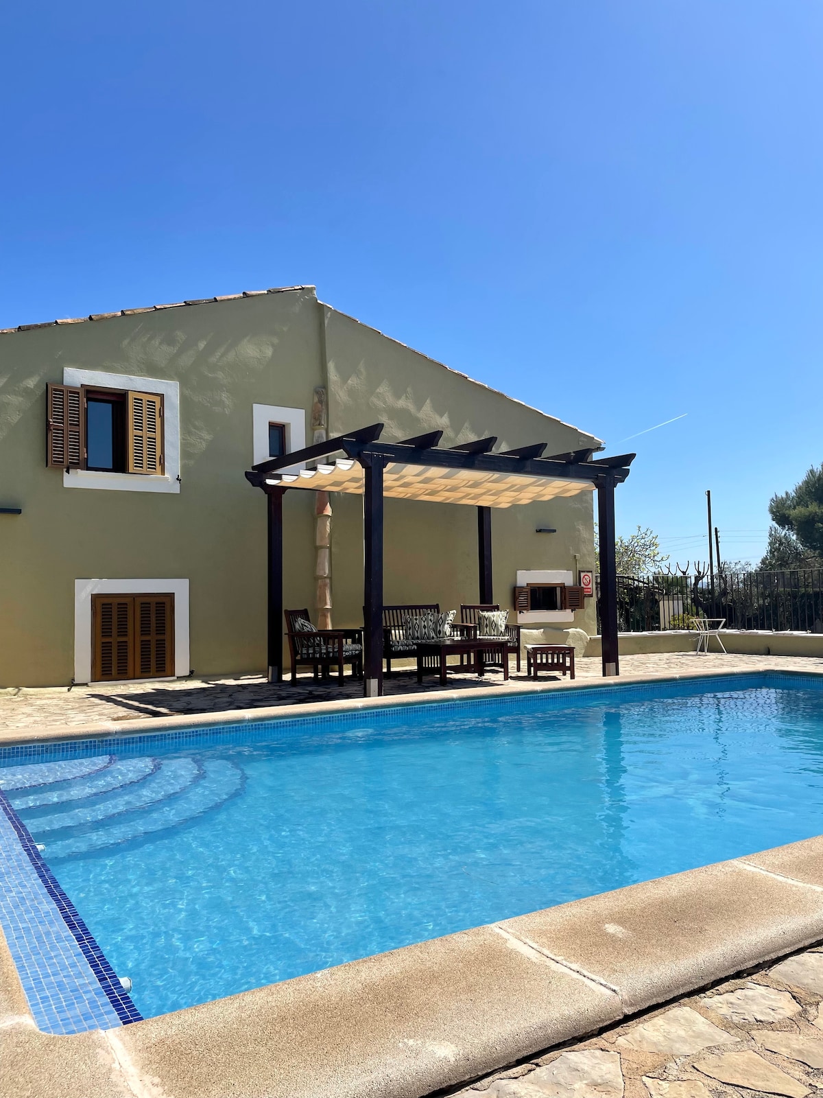 A two-story house is highlighted by a covered terrace with wooden furniture, overlooking a refreshing pool. Sunlight reflects off the water, while shutters frame the windows, contributing to the rustic charm. A clear blue sky provides a serene backdrop.