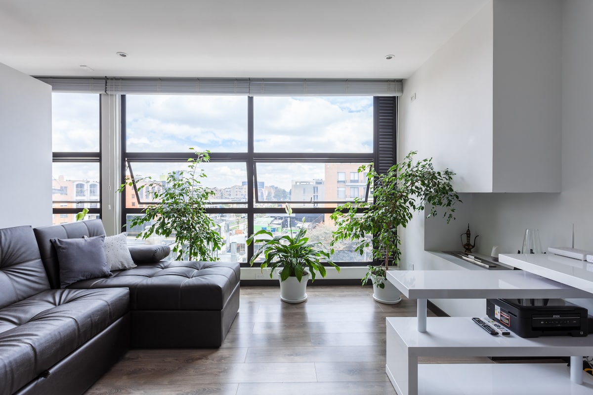 A spacious living area is highlighted by large windows that allow natural light to fill the space. A dark sectional sofa is accompanied by decorative throw pillows, while potted plants add a touch of greenery. A minimalist white desk is positioned to one side.