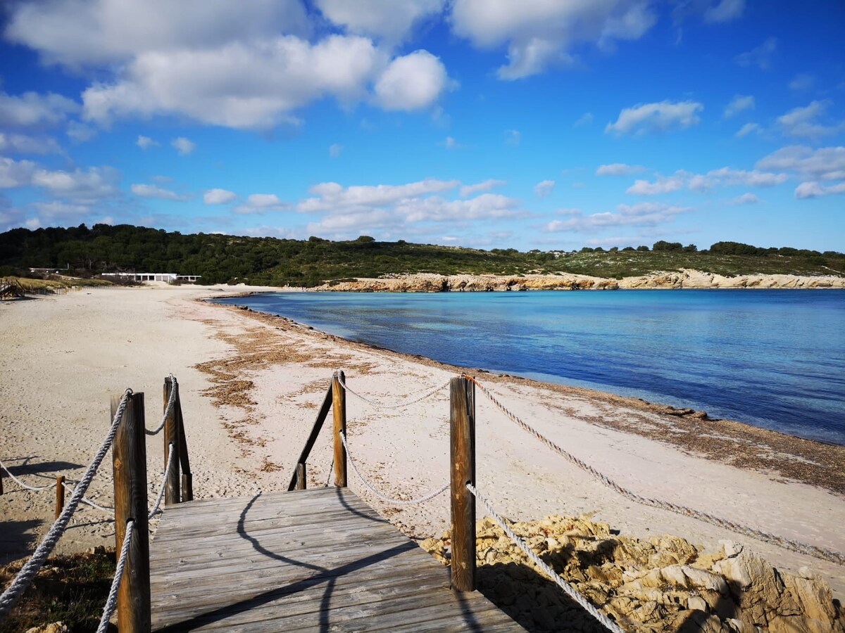 A wooden pathway leads to a sandy beach, complemented by calm blue waters. Sunlit clouds dot the sky above, and green landscapes can be seen in the distance. This serene coastal view offers a peaceful atmosphere, ideal for relaxation.