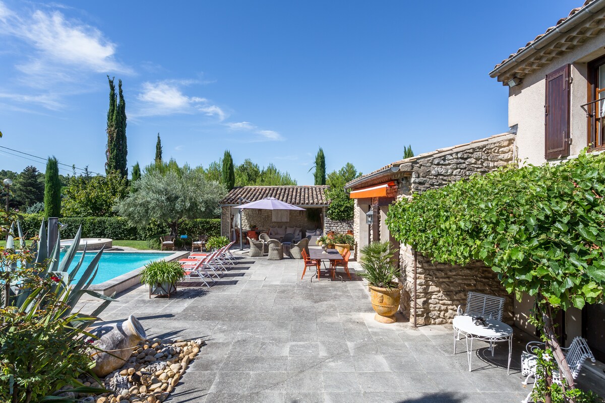 An outdoor area features a large swimming pool surrounded by sun loungers and a shaded seating space. Lush greenery and flowering plants enhance the tranquil ambiance. Terracotta pots and stone paths add to the rustic charm of the patio adjoining the house.