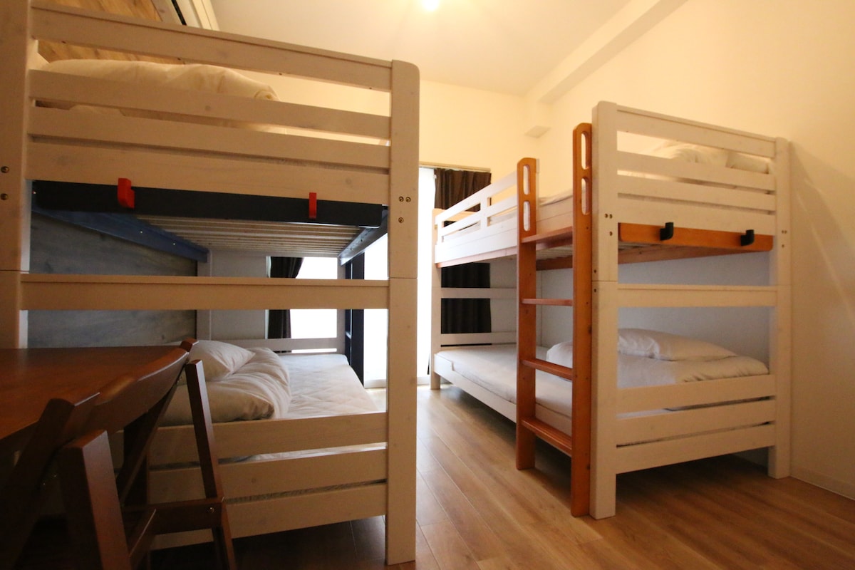 The room features two sets of bunk beds, accommodating up to four guests. A modest table and chairs are also included, creating a functional space for group gatherings. Natural light enters through a nearby window, complementing the warm wooden flooring.