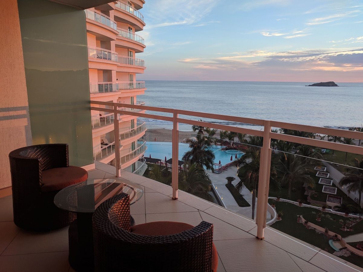 A balcony features two woven chairs and a glass table, offering a view of the tranquil sea and the infinity pool below. The distant island is visible against a colorful sky during sunset, creating a serene environment surrounded by lush landscaping.