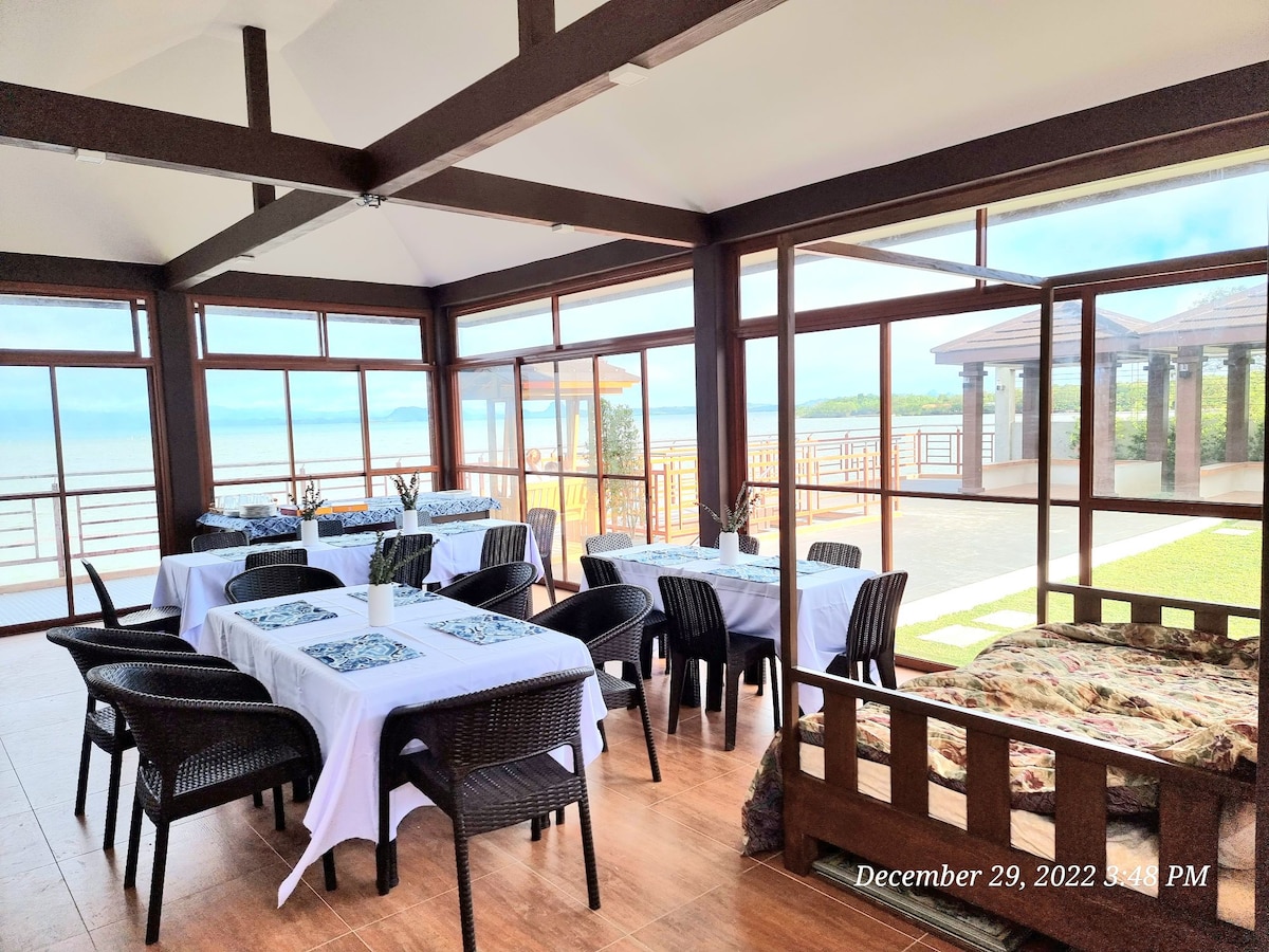A dining area is presented with several tables set for meals, featuring white tablecloths. Large windows allow abundant natural light and offer views of Pagbilao Bay. Outdoor seating can be seen through the glass doors, creating a seamless connection between indoor and outdoor spaces.