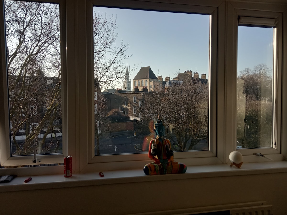A view from a large window displays the surrounding street and buildings of old London, framed by clear glass. A colorful statue sits on the sill, adding an artistic touch, while trees with bare branches suggest the season.