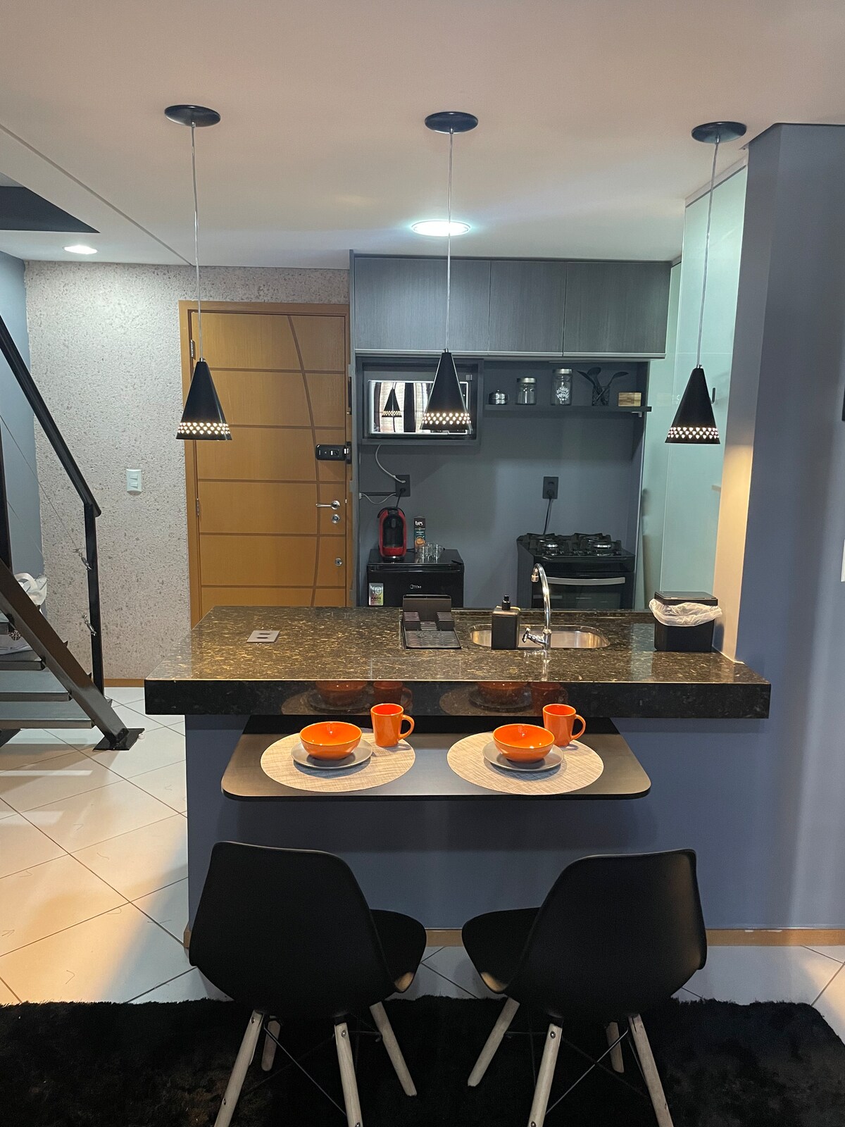 A modern kitchen area features a dark granite countertop with two orange cups and plates set at a small dining table. Hanging black pendant lights illuminate the space. Cabinets and appliances are neatly arranged in the background, creating a functional and inviting atmosphere.
