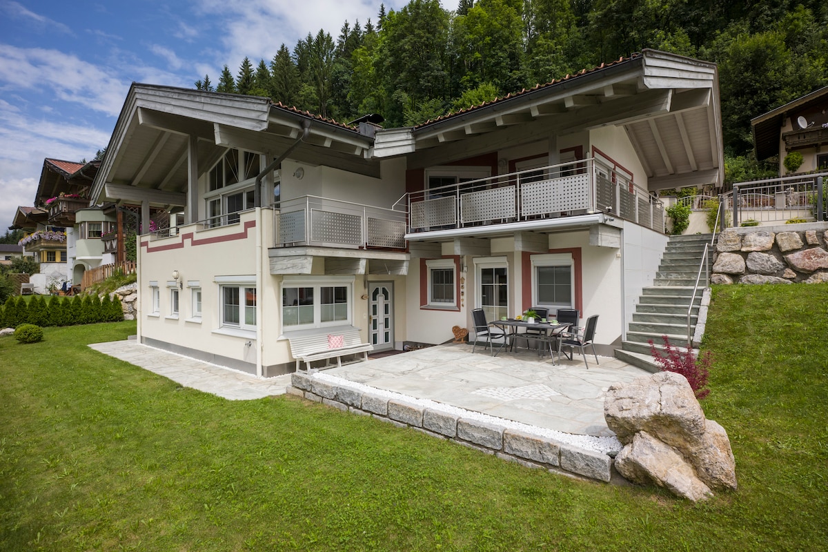 The exterior of a modern apartment is displayed, featuring a spacious patio with a stone pathway and outdoor dining area. The well-maintained lawn and surrounding trees contribute to a serene environment. The structure consists of two levels, with a balcony offering additional outdoor space.