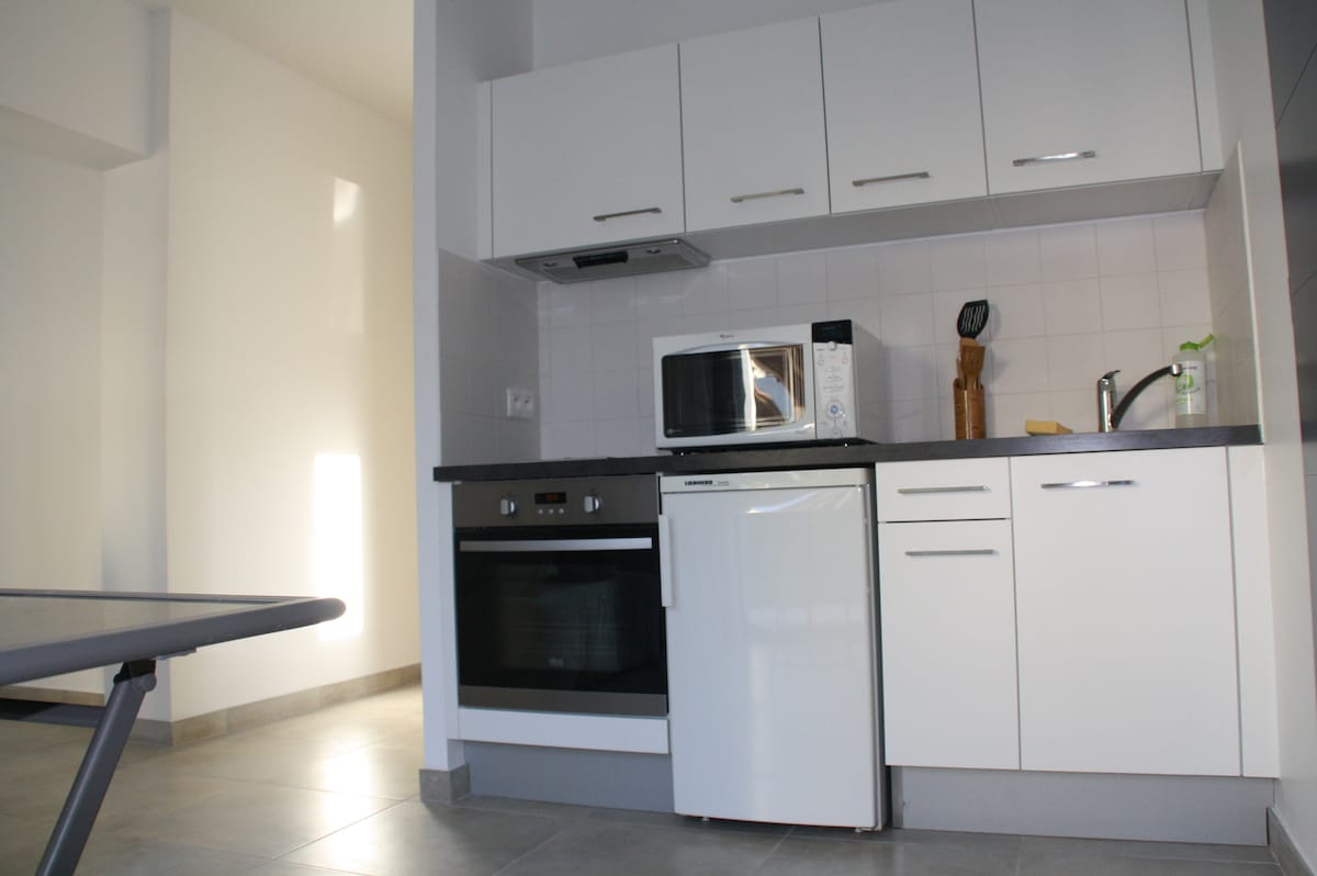 A modern kitchen is presented with sleek white cabinetry and gray countertops. Appliances include a microwave, oven, and refrigerator, complemented by a wooden utensil holder. Natural light floods the space, highlighting the tile flooring and minimalist design.