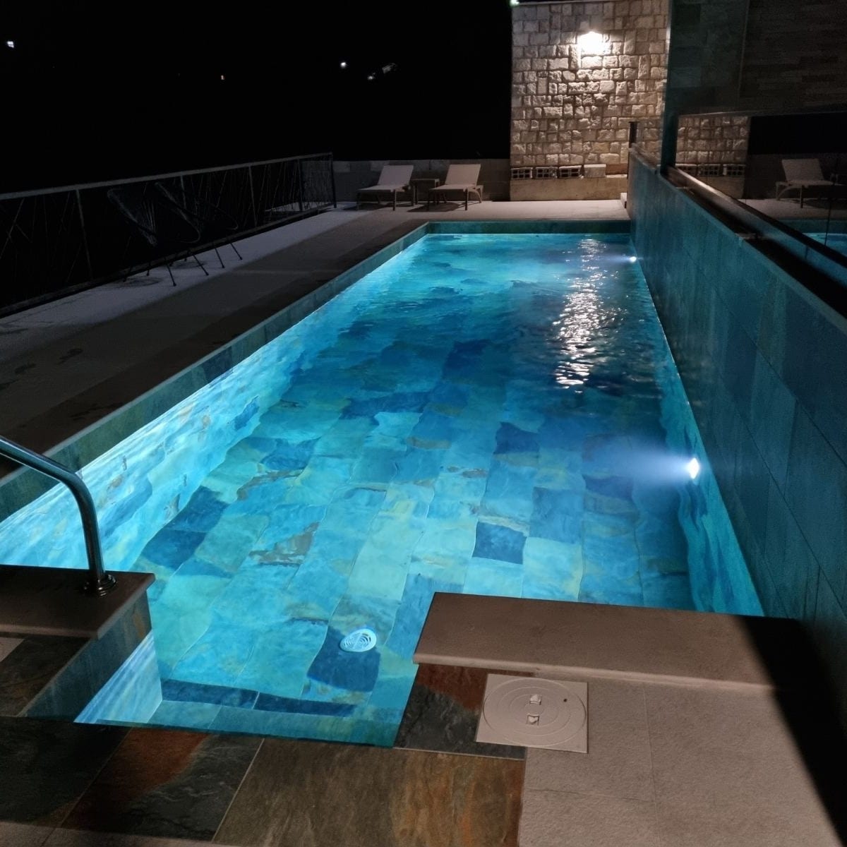 A serene swimming pool is illuminated at night, featuring cool blue tiles that reflect the light. The pool area is bordered by a stone wall, and several lounge chairs are positioned nearby, creating a restful atmosphere for relaxation.