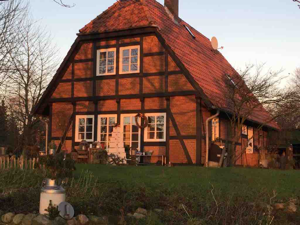 The charming half-timbered house features a warm brick facade, complemented by a sloped red roof. Large windows are framed with white paint, and decorative elements like a wreath adorn the entrance. An inviting outdoor space surrounds the property, enhancing its rural appeal.