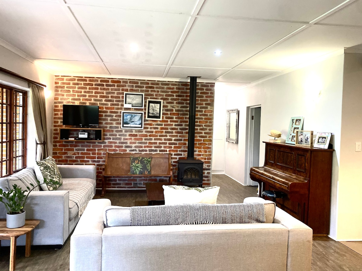 A spacious living area features a cozy seating arrangement with light grey sofas and a wooden coffee table. A brick wall serves as a backdrop for a television, while a small indoor plant adds a touch of greenery. A piano is positioned adjacent to a large window, enhancing the room's charm.