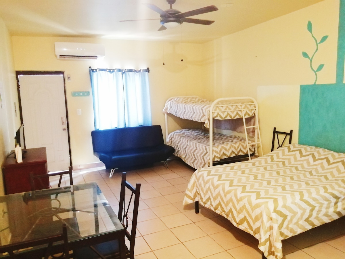 A spacious loft area is presented, featuring a large bed and a bunk bed with a chevron pattern. A glass dining table and chairs occupy one section, while a ceiling fan provides ventilation. Natural light filters through a window covered with light curtains.