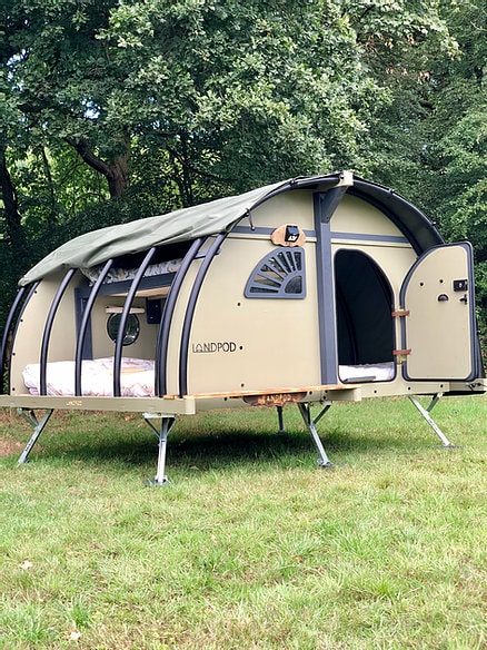 The LandPod is situated on a grassy area, featuring a sturdy frame and a curved roof. The entrance is partially open, revealing a cozy interior with a double bed and a bunk bed. Natural surroundings add to the outdoor experience.