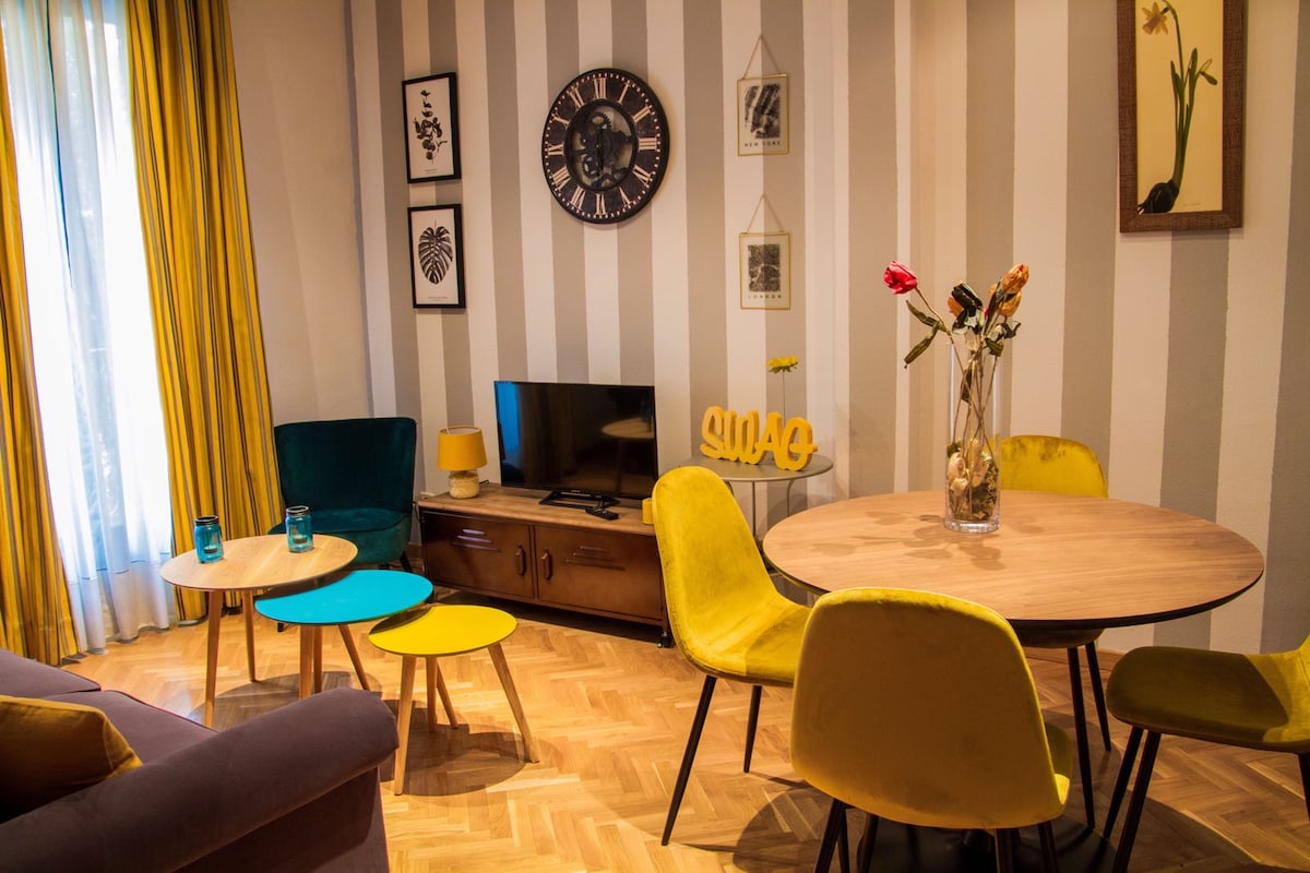 A bright living area features a round wooden dining table surrounded by colorful chairs in yellow and green. A wall-mounted television is positioned above a low wooden cabinet. The space is enhanced by tall windows with flowing curtains and decorative wall art.