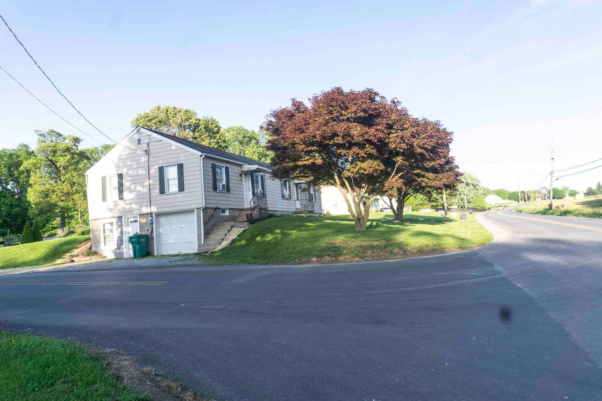 A two-story townhouse is positioned on a grassy corner lot, surrounded by mature trees and shrubbery. The facade features light-colored siding, and steps lead to the main entrance. The road curves gently around the property, framed by a clear blue sky.