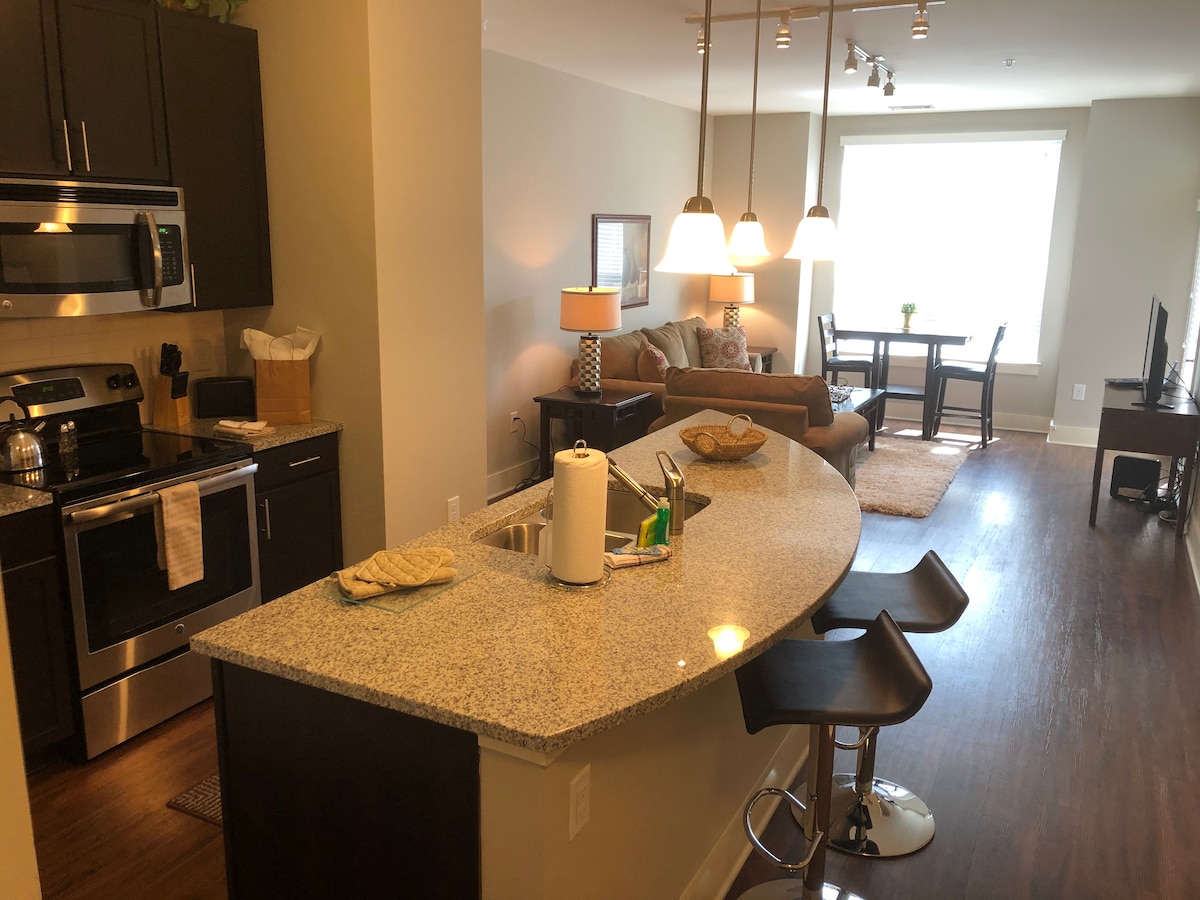 An open-concept kitchen and living area are presented with modern fixtures. The kitchen features a curved countertop with bar stools, stainless steel appliances, and ample cabinetry. In the background, a seating area includes a sofa and a dining table near a large window allowing natural light.