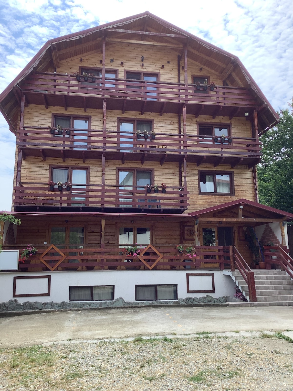 A multi-story wooden chalet is presented, featuring a traditional design with multiple balconies adorned with flower pots. Large windows allow natural light to illuminate the interior. A set of stairs leads to the welcoming entryway, enhancing accessibility to the main level.