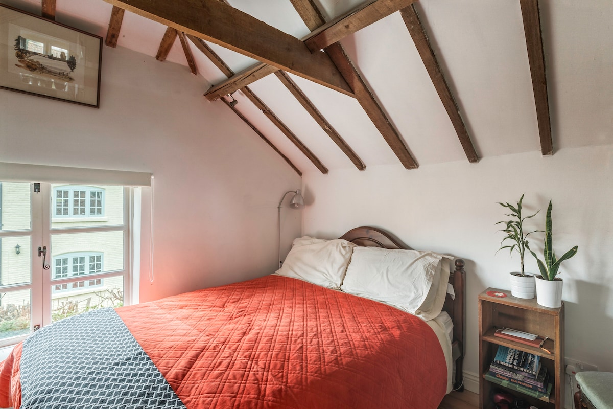 A compact sleeping area features a spacious bed adorned with an orange quilt. Natural light enters through a nearby window, highlighting exposed wooden beams on the ceiling. A small bedside table with a plant completes the cozy atmosphere.
