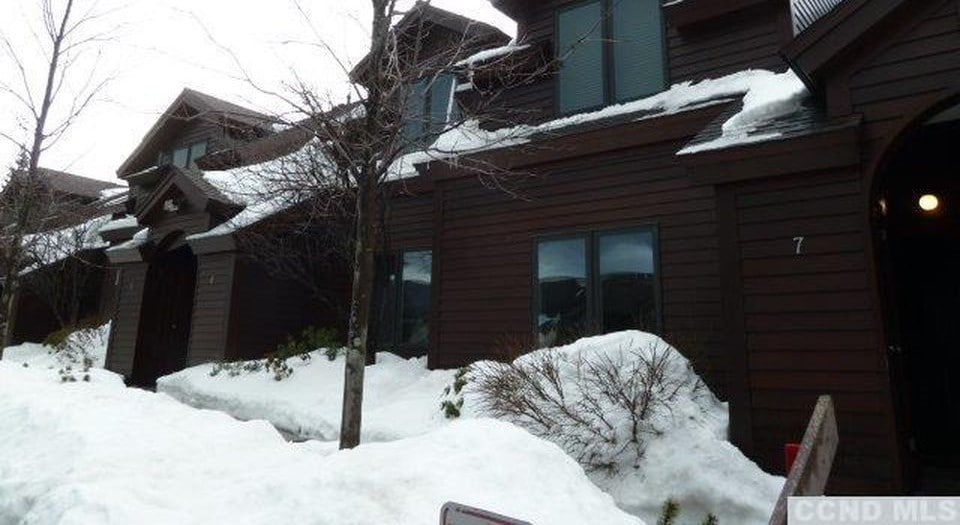 A dark wooden exterior with multiple units is presented, surrounded by snow-covered ground and sparse greenery. Large windows allow for natural light, while a sloped roof retains a blanket of snow. The entrance is framed by trees, enhancing the winter scene.