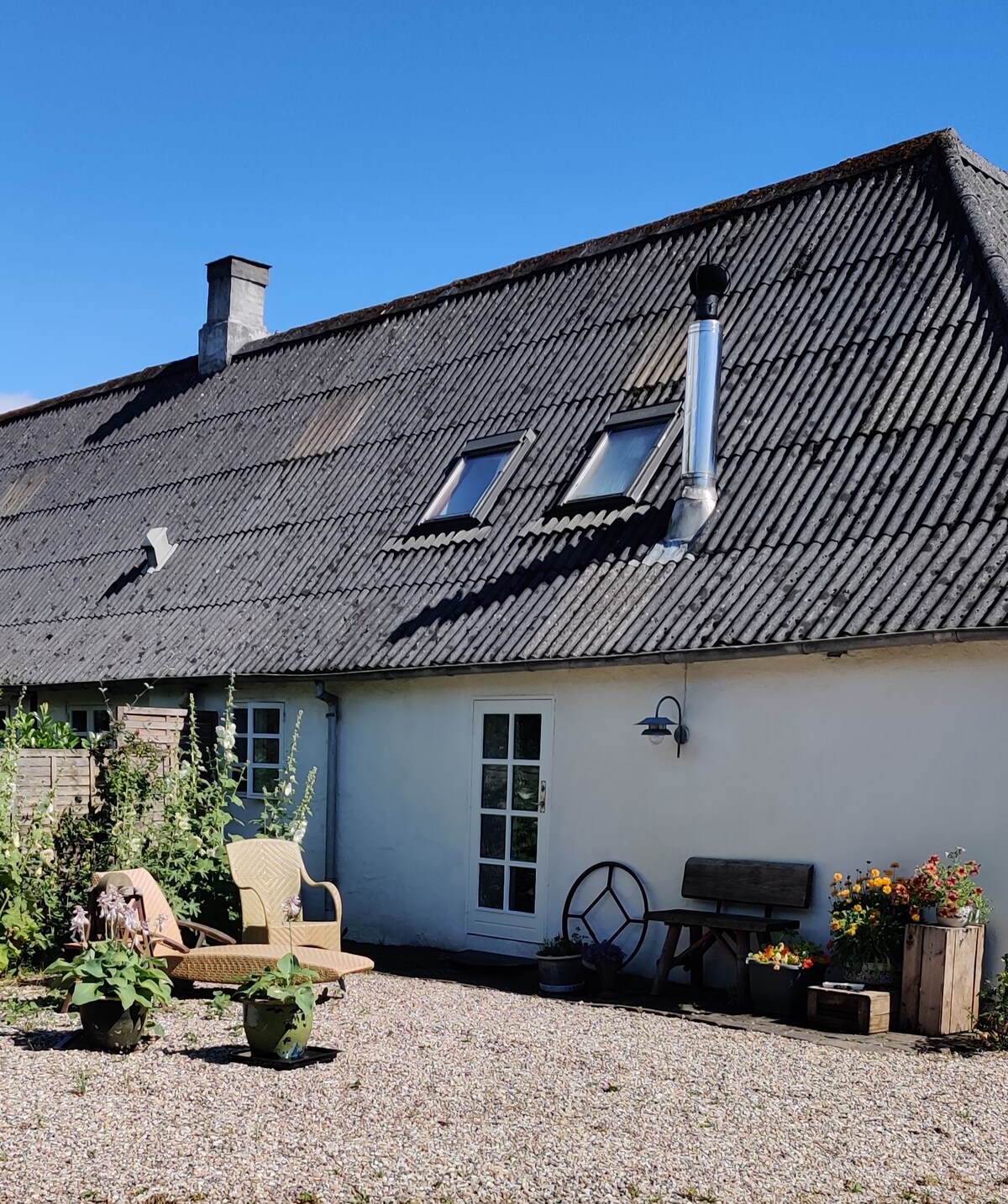 The exterior of a charming building displays a sloped, thatched roof with skylights. A small seating area features a wicker chair and rustic table, surrounded by flower pots and natural greenery, providing a serene outdoor space perfect for enjoying the surrounding landscape.