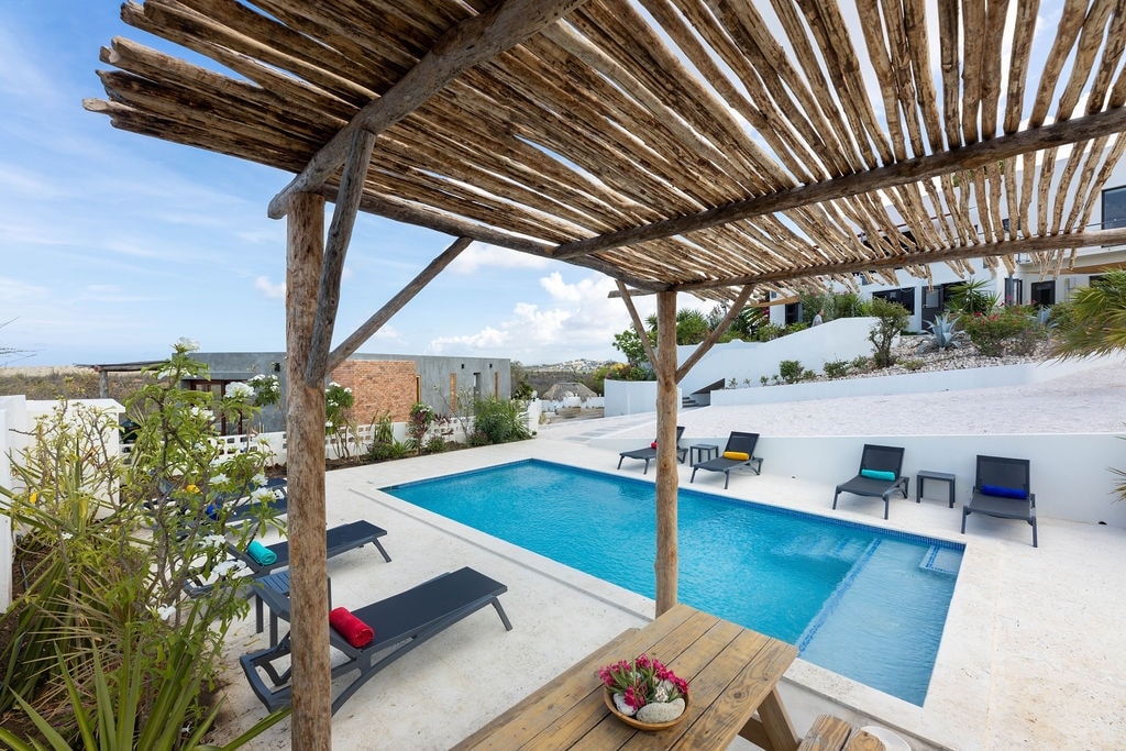 An inviting outdoor space is showcased, featuring a rectangular swimming pool surrounded by sun loungers in various colors. A shaded wooden pergola offers seating with a wooden table adorned with a bowl of fruit. Lush greenery frames the area against a clear blue sky.