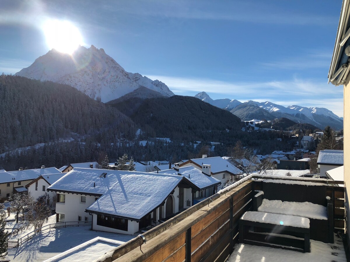 A panoramic view is presented from the sunny balcony, showcasing snow-covered mountains and the surrounding village. The sunlight casts a warm glow over the landscape, highlighting the peaks and gently sloping hills, while the wooden railing offers an inviting frame to the scenic backdrop.