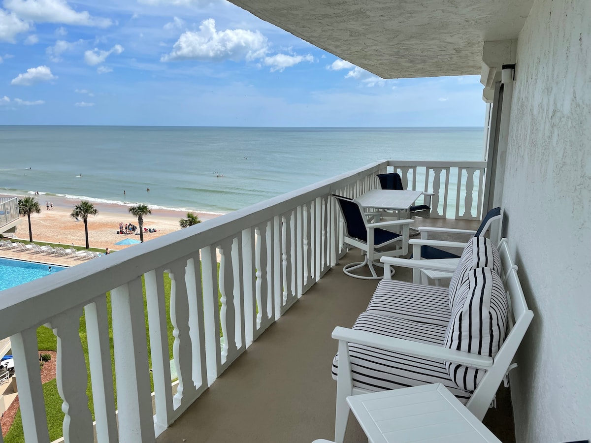 A spacious balcony features white outdoor furniture, including striped and solid blue cushioned chairs and a table. The serene ocean view stretches towards the horizon, with a sandy beach and palm trees visible below under a bright blue sky.