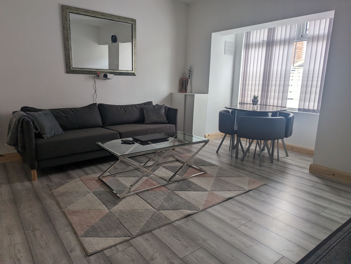A modern living area features a dark grey sofa positioned near a glass coffee table on a patterned rug. Natural light filters through wide windows with sheer vertical blinds, illuminating a small dining table set for four. Light wood flooring enhances the space.