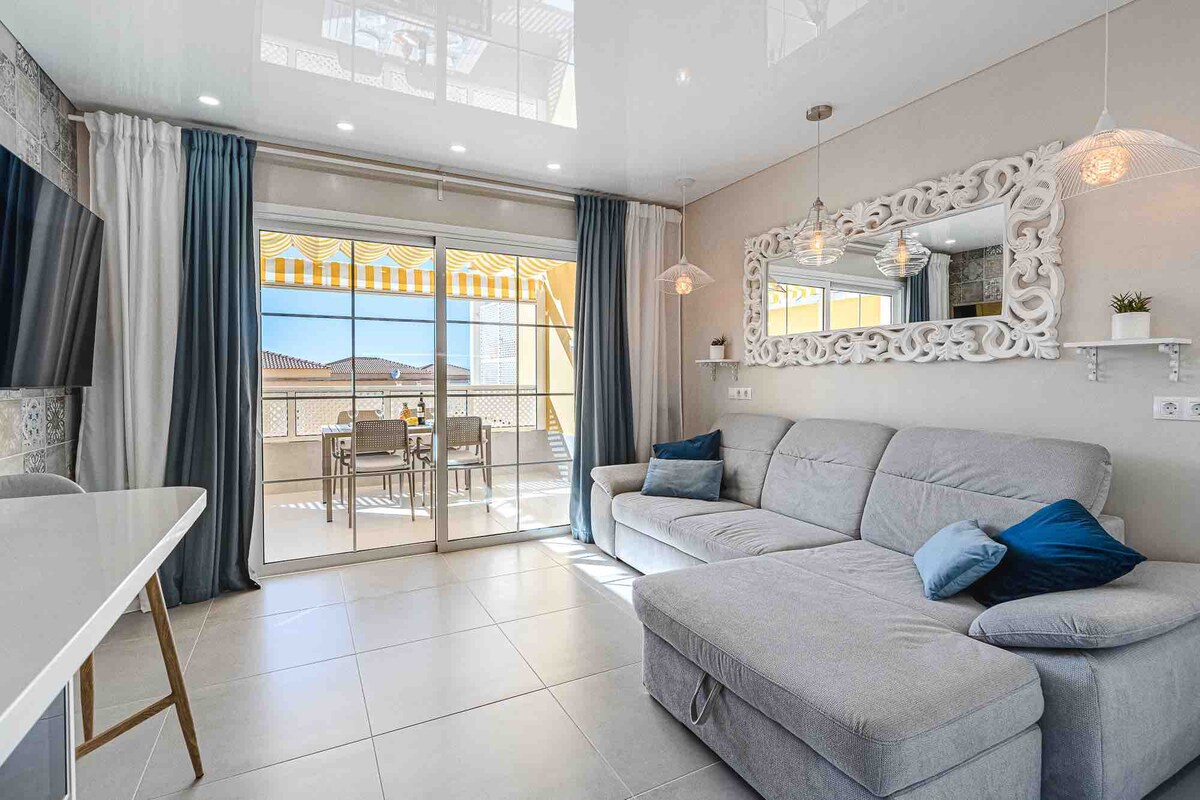 A modern living area features a light gray sectional sofa with blue accent pillows facing a wall-mounted television. Large sliding glass doors provide access to a private terrace, where a dining set can be seen. Neutral tones and pendant lights add to the spacious ambiance.