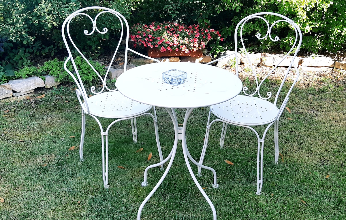 A small outdoor table is set with two white metal chairs. The table features a circular shape and a dotted design. A glass ashtray sits at the center. A garden of blooming flowers serves as a backdrop, adding a splash of color to the greenery.