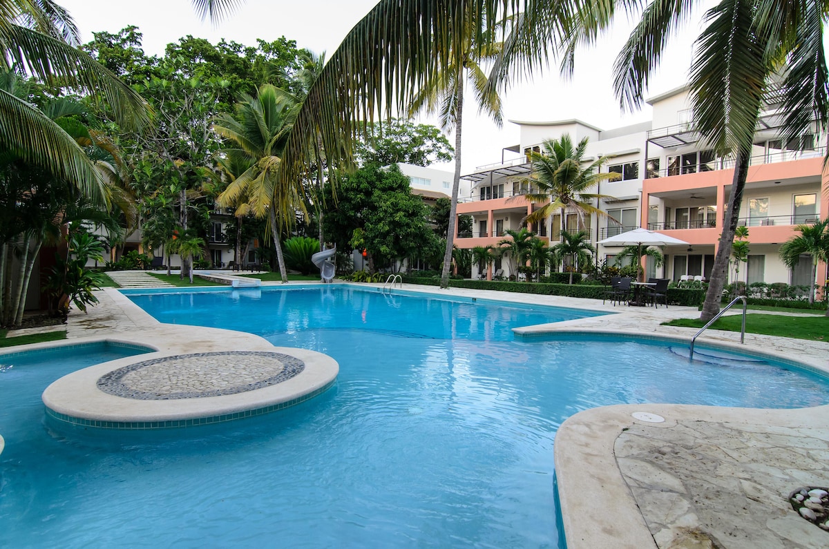 A serene swimming pool is surrounded by lush tropical landscaping, featuring palm trees and well-maintained gardens. The pool's gentle curves invite relaxation, with lounge chairs positioned nearby under shaded areas. The modern condominium buildings are visible in the background, creating a harmonious atmosphere.