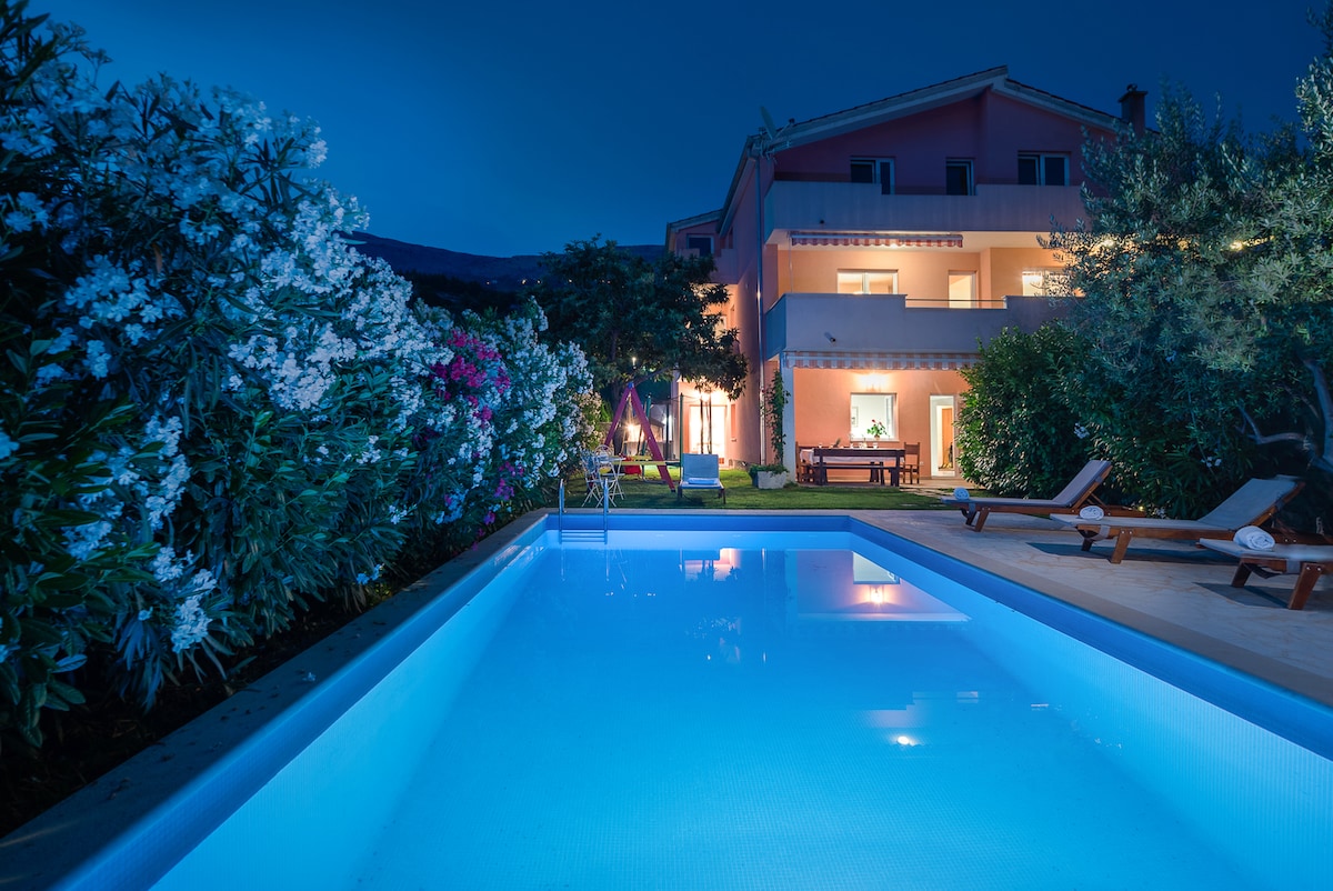 A serene evening view of the villa shows a well-lit swimming pool surrounded by lush greenery and blooming flowers. The two-story villa, painted in soft pink, stands in the background, with sun loungers positioned nearby, inviting relaxation amidst the tranquil setting.