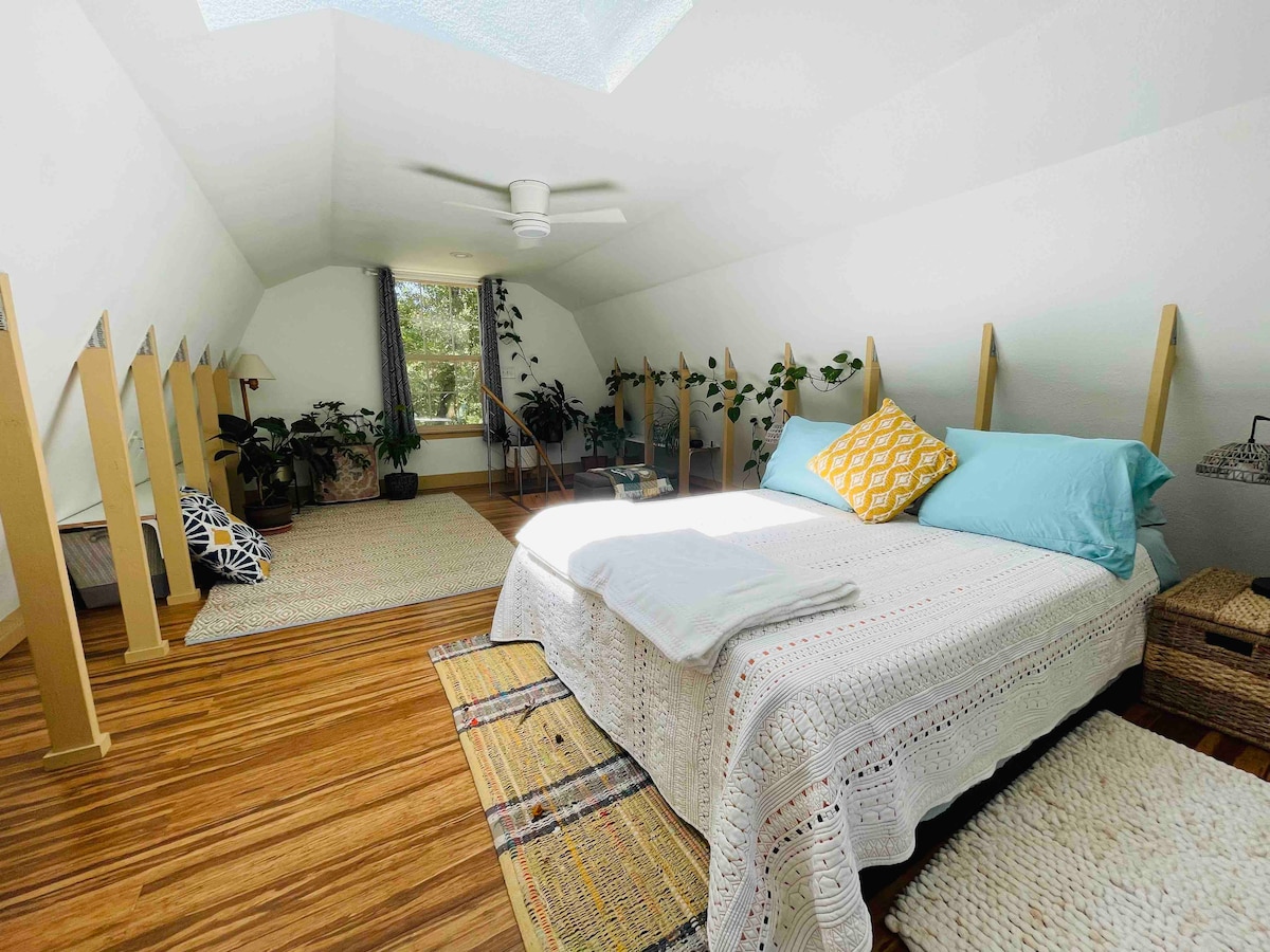 A spacious loft bedroom is filled with natural light from a skylight above. A cozy bed is adorned with soft blue and white linens. Potted plants are arranged around the room, and light wooden accents create a warm atmosphere.