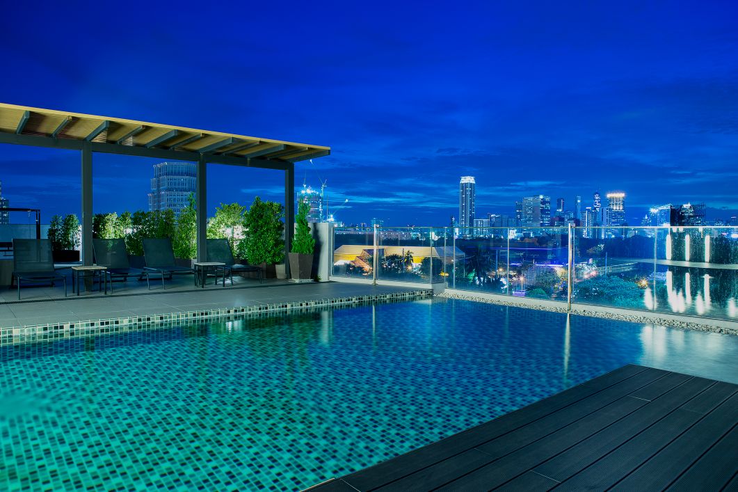 A rooftop pool is presented, featuring a modern design with turquoise tiles. Surrounding seating areas provide views of the city skyline. The clear night sky enhances the serene atmosphere, while the city lights create a vibrant backdrop.