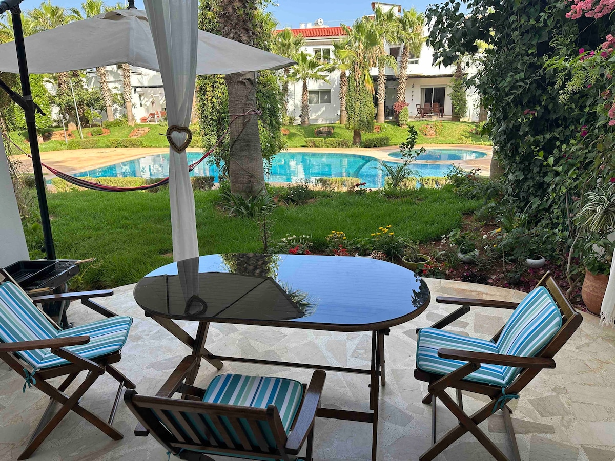 An outdoor seating area is featured with a black table and striped chairs, set against a backdrop of lush greenery and a shared swimming pool. Palm trees and flowering plants surround the space, creating a calm environment for relaxation.