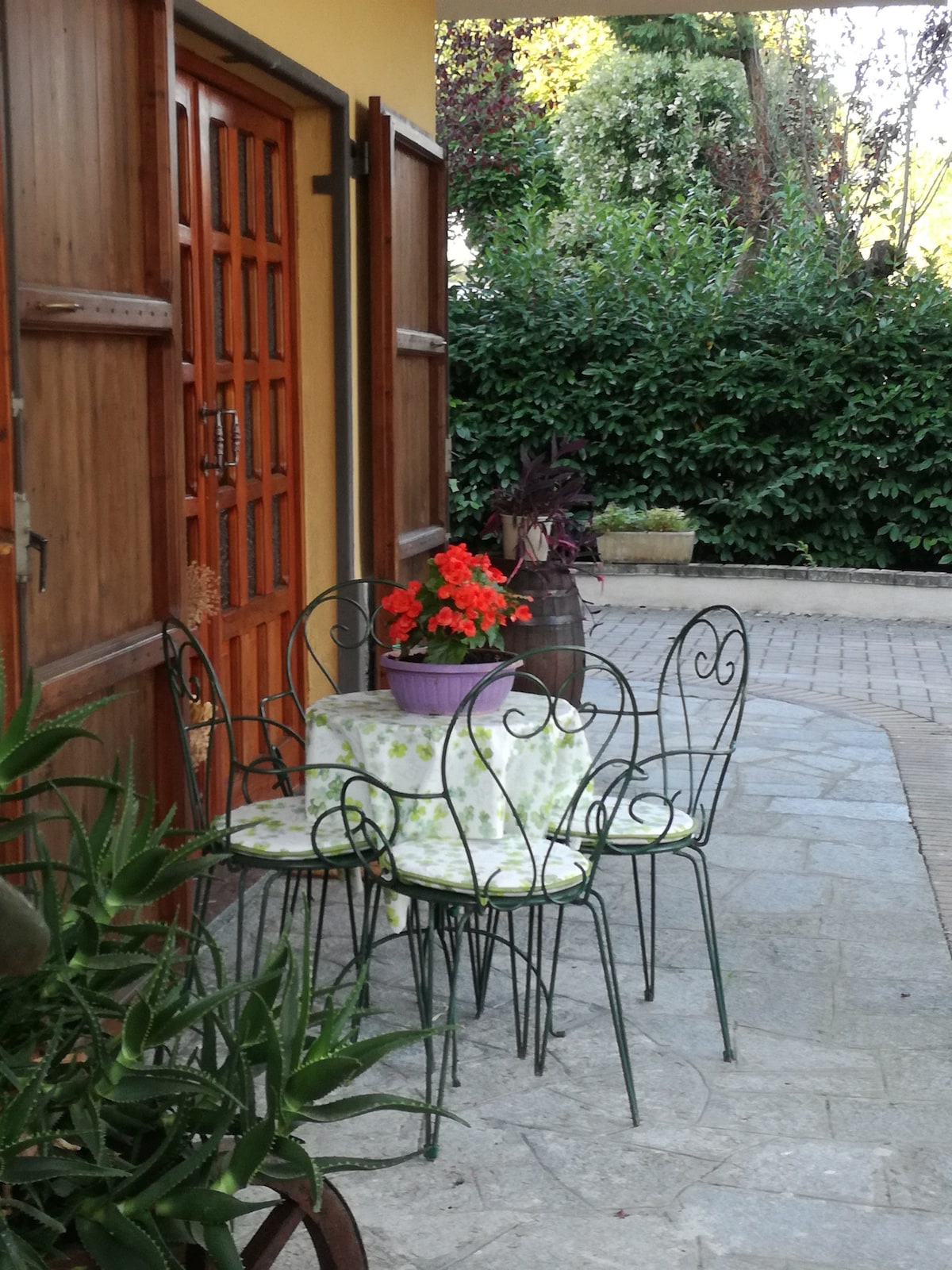 A small outdoor seating area is visible, featuring a round table surrounded by four iron chairs, each with patterned cushions. A pot of red flowers adds a touch of color, while lush greenery creates a serene backdrop.