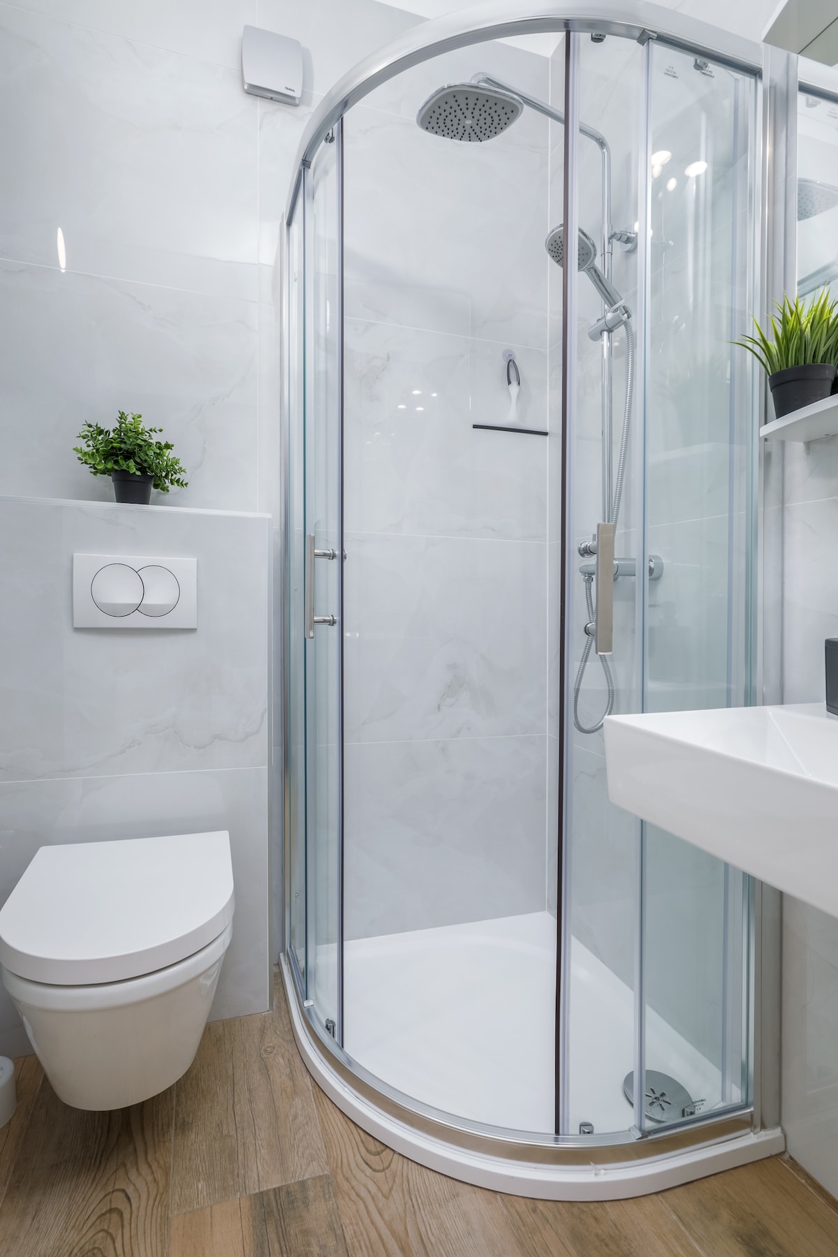 A modern bathroom is presented, featuring a curved glass shower enclosure and a minimalist toilet. Soft greenery is visible on the wall shelf, alongside elegant gray finishes and wooden flooring that contribute to the clean design.