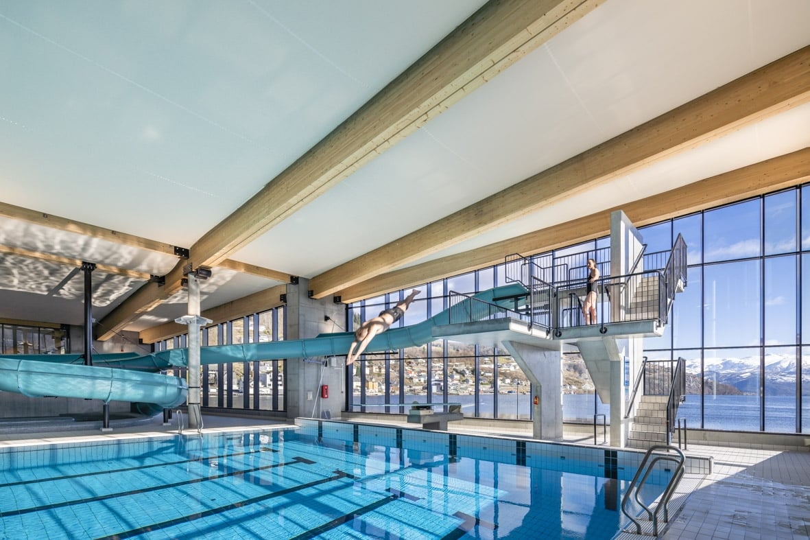 An indoor swimming pool is highlighted by large windows that offer views of the surrounding landscape. Two diving boards are positioned above the water, with a slide curving into the pool area. Sunlight illuminates the space, enhancing its spacious and modern design.